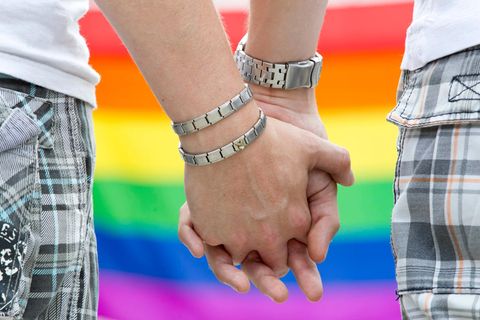 Die Hände zweier Männer, ineinander verschränkt, vor Regenbogenfahne