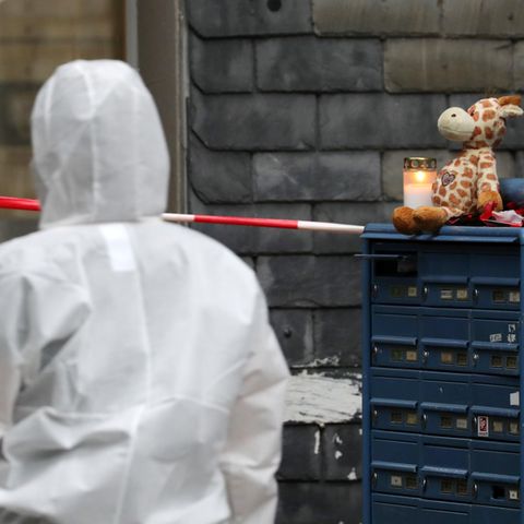 Eine Person in weißem Overall geht auf einen Hauseingang zu. Rechts von ihr sind blaue Briefkästen, auf denen eine Kerze brennt