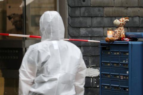 Eine Person in weißem Overall geht auf einen Hauseingang zu. Rechts von ihr sind blaue Briefkästen, auf denen eine Kerze brennt