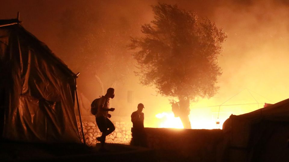 Feuer ist im Flüchtlingslager Lesbos zu sehen, vorn gehen zwei Personen