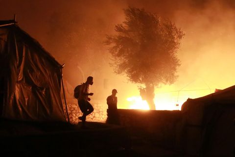 Feuer ist im Flüchtlingslager Lesbos zu sehen, vorn gehen zwei Personen