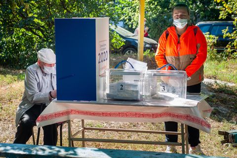 Russland, Tambow: Ein Mann mit Mundschutz füllt hinter einer Trennwand seinen Stimmzettel aus. Mitglieder der Wahlkommission der Bezirke warten im Freien in einem Wahllokal auf die Wähler.