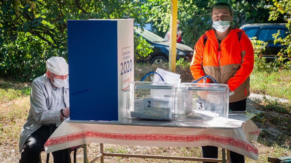 Russland, Tambow: Ein Mann mit Mundschutz füllt hinter einer Trennwand seinen Stimmzettel aus. Mitglieder der Wahlkommission der Bezirke warten im Freien in einem Wahllokal auf die Wähler.