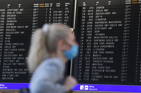 Frau mit Mundschutz steht im Terminal 1 des Frankfurter Flughafens in der Abflughalle vor einer großen Anzeigetafel