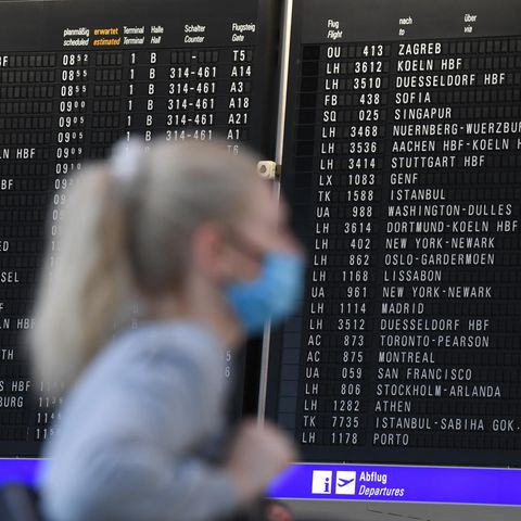 Frau mit Mundschutz steht im Terminal 1 des Frankfurter Flughafens in der Abflughalle vor einer großen Anzeigetafel