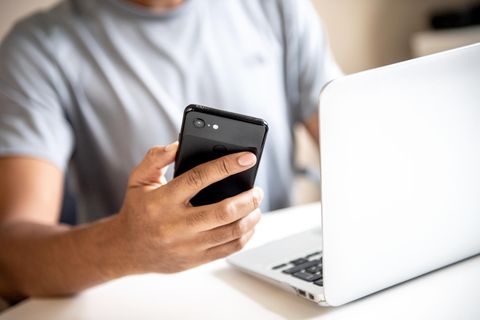 Wlan Passwort anzeigen: Junger Mann sitzt mit Smartphone in der Hand vor einem Laptop
