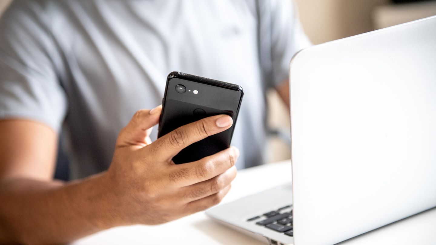 Wlan Passwort anzeigen: Junger Mann sitzt mit Smartphone in der Hand vor einem Laptop
