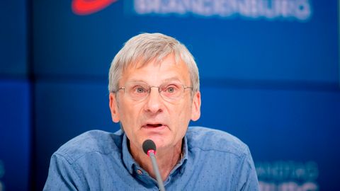 Ein Mann mit grauem Seitenscheitel und randloser Brille sitzt auf einem blauen Podium und spricht in ein Mikrofon