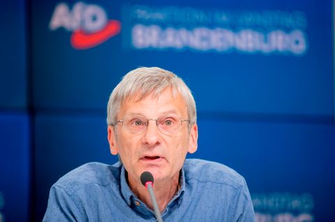Ein Mann mit grauem Seitenscheitel und randloser Brille sitzt auf einem blauen Podium und spricht in ein Mikrofon