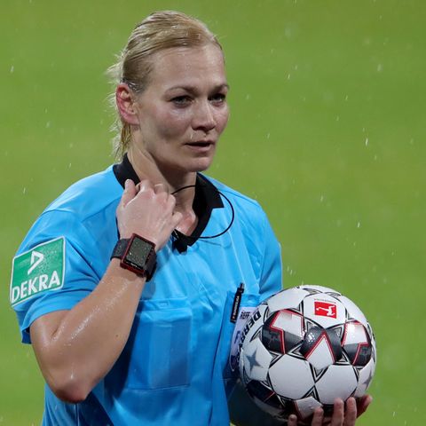 Bibiana Steinhaus steht im hellblauen Schiedsrichter-Trikot im Regen auf dem Rasen und hält einen Ball im linken Arm