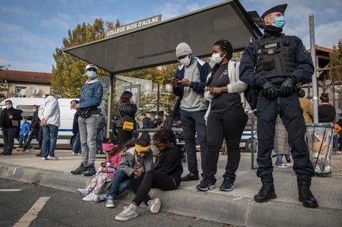 Am Dach eines Bushaltestellen-Häuschens steht "College Bois d'Aulne". Vor dem Häuschen warten Schüler und ein Polizist