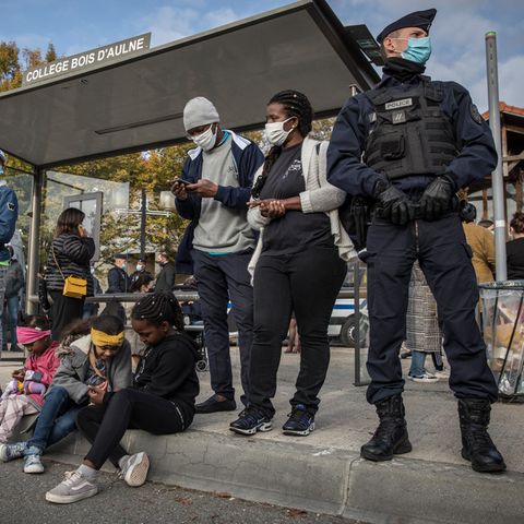 Am Dach eines Bushaltestellen-Häuschens steht "College Bois d'Aulne". Vor dem Häuschen warten Schüler und ein Polizist