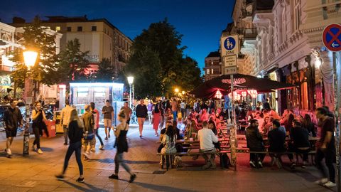Auf einem breiten Fußweg sitzen zur blauen Stunde junge Menschen auf Bierbänken und trinken trotz Corona