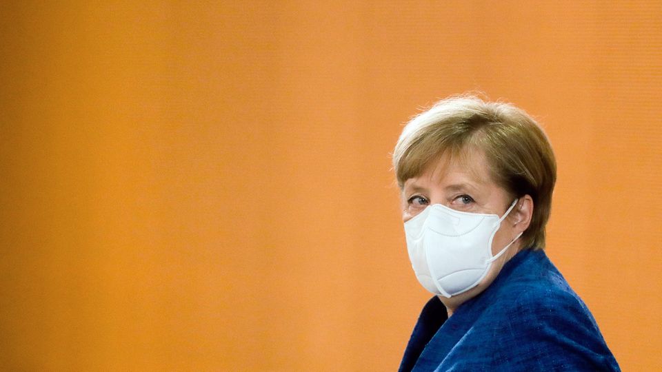 Bundeskanzlerin Angela Merkel trägt einen weißen Mund-Nasen-Schutz sowie ein blauen Blazer. Sie schaut über die linke Schulter