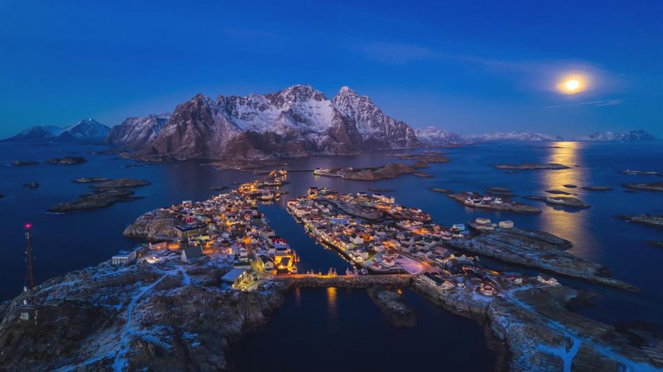 Norwegen, Lofoten: Henningsvær  Im Schein des Mondes leuchten die roten Fischerhäuser und die gelblichen Felsen in warmen Tonen, während der umgebende Ozean ein tiefes Blau annimmt. 