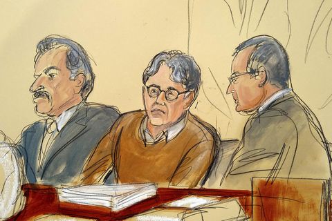 Ein Mann mit Brille und braunem Pullover sitzt zwischen zwei Anwälten in Anzügen