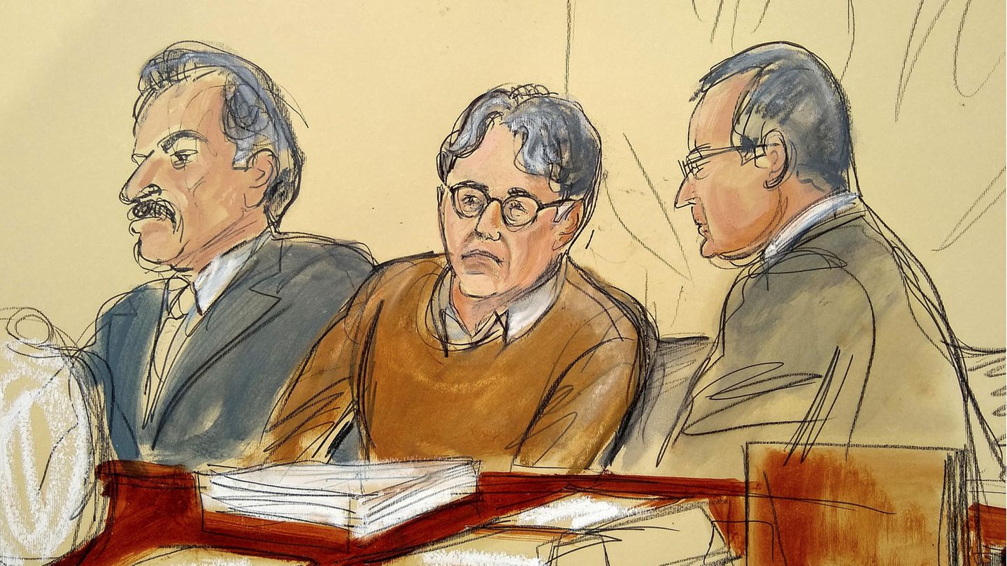 Ein Mann mit Brille und braunem Pullover sitzt zwischen zwei Anwälten in Anzügen