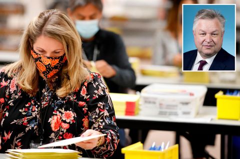 Eine blonde Frau mit Mundschutz und geblümter Bluse sitzt an einem Tisch und sortiert Briefwahl-Unterlagen