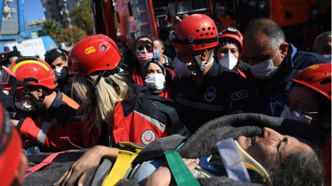 Auf einer Liege tragen Retter in roten Jacken und Helmen eine schwarzhaarige Frau über die Trümmer eines eingestürzten Gebäudes