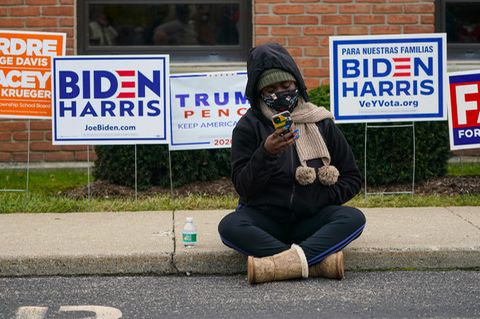 Eine Frau sitzt mit ihrem Handy auf dem Bürgersteig, hinter ihr sind Schilder für Donald Trump oder Joe Biden.