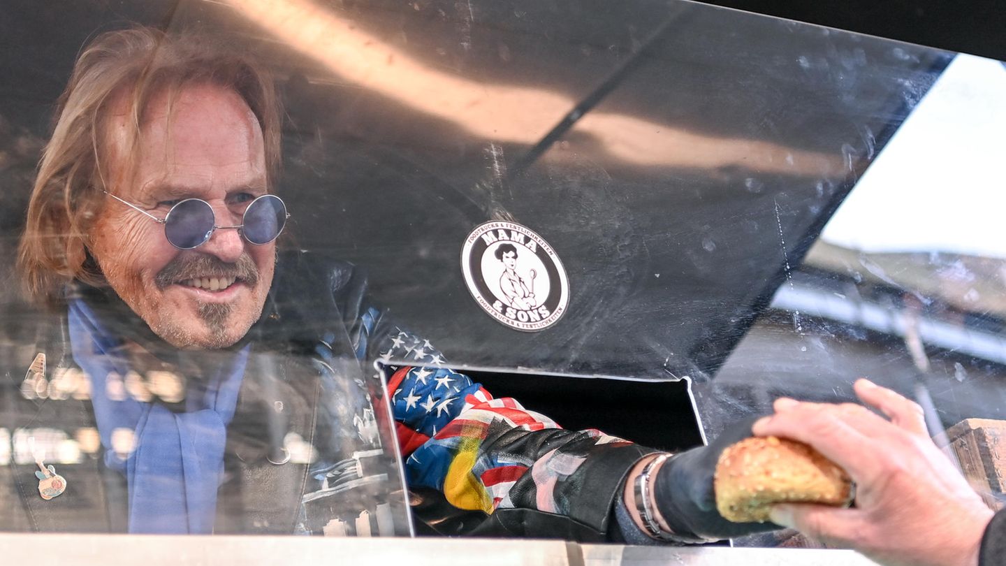 Frank Zander reicht durch eine Öffnung in einer Plexiglasscheibe ein Brötchen an