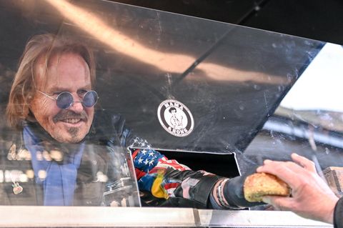 Frank Zander reicht durch eine Öffnung in einer Plexiglasscheibe ein Brötchen an