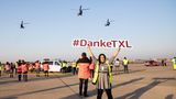 Auf dem Vorfeld hält eine Frau ein Schild mit dem Hashtag "Danke-TXL" in die Luft, während im Hintergrund Hubschrauber der Bundespolizei zum Abschied vorbeifliegen.