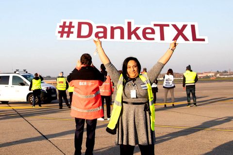 Auf dem Vorfeld hält eine Frau ein Schild mit dem Hashtag "Danke-TXL" in die Luft, während im Hintergrund Hubschrauber der Bundespolizei zum Abschied vorbeifliegen.