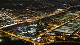 Wolfsburg: Volkswagenwerk und Autostadt  Rechts leuchten die Hallen des Automobilwerkes mit dem Freizeitpark, der Autostadt weiter links. In den beiden Doppeltürmen warten neue Fahrzeuge auf die Auslieferung an Kunden, links daneben steht das kreisrunde Ritz-Carlton-Hotel. Auf der anderen Seite des dunklen Mittellandkanals liegt die Stadt mit dem Science Center Phæno am linken Bildrand.