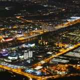 Wolfsburg: Volkswagenwerk und Autostadt  Rechts leuchten die Hallen des Automobilwerkes mit dem Freizeitpark, der Autostadt weiter links. In den beiden Doppeltürmen warten neue Fahrzeuge auf die Auslieferung an Kunden, links daneben steht das kreisrunde Ritz-Carlton-Hotel. Auf der anderen Seite des dunklen Mittellandkanals liegt die Stadt mit dem Science Center Phæno am linken Bildrand.