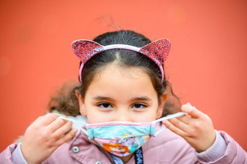 Eine Grundschülerin mit langen, braunen Locken setzt sich eine hellblaue Maske auf. Sie trägt einen Haarreif mit Glitzer-Ohren