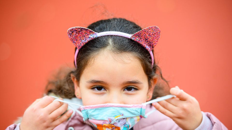 Eine Grundschülerin mit langen, braunen Locken setzt sich eine hellblaue Maske auf. Sie trägt einen Haarreif mit Glitzer-Ohren