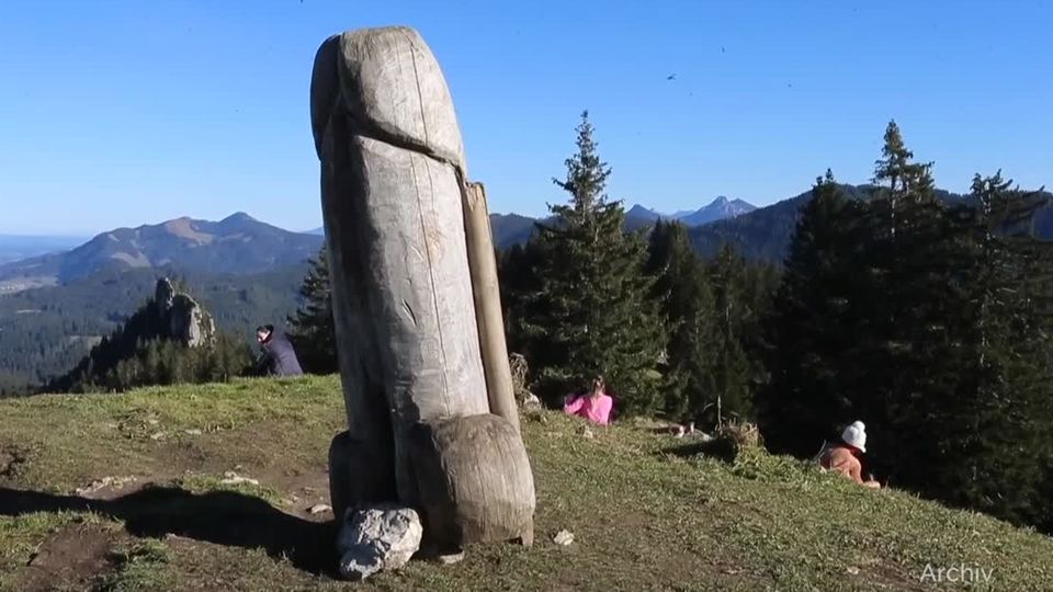 Penis aus Holz im Allgäu Auf einer Bergkuppe steht ein verwitterter, erigierter Penis aus Holz. Dahinter rasten Wanderer und genießen den Ausblick