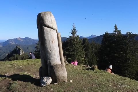 Auf einer Bergkuppe steht ein verwitterter, erigierter Penis aus Holz. Dahinter rasten Wanderer und genießen den Ausblick