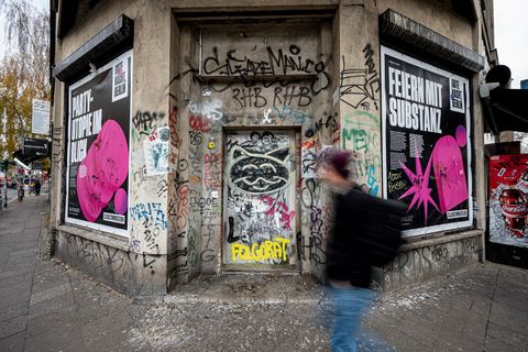 Eine braune Häuserecke in Berlin, darauf sind Poster über das Feiern. Ein Mann läuft auf dem Bild vorbei.