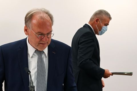 Ein Mann mit Brille und Halbglatze steht im Anzug im Vordergrund, während hinter ihm ein anderer Mann seitlich weggeht
