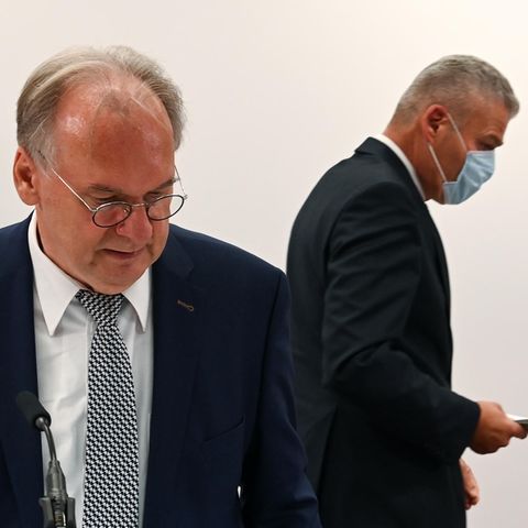 Ein Mann mit Brille und Halbglatze steht im Anzug im Vordergrund, während hinter ihm ein anderer Mann seitlich weggeht