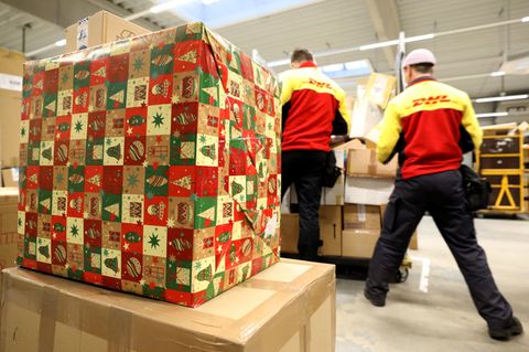 Ein großes Paket in Weihnachtspapier liegt auf einem Stapel Kartons, während im Hintergrund DHL-Mitarbeiter