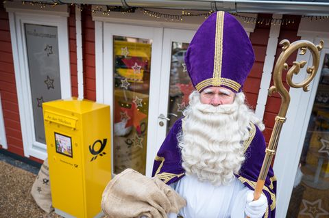Ein Mann mit weißem Vollbart, violettem Bischofstab steht vor einem gelben Briefkasten