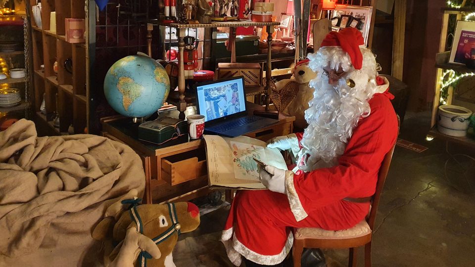 Ein Mann in einem Weihnachtsmann-Kostüm sitzt mit Smartphone und Tablet an einem kleinen Schreibtisch
