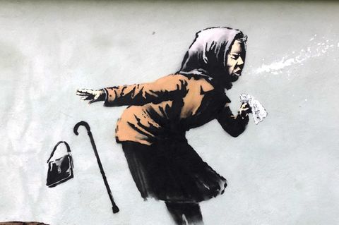 Auf einer hellgrünen Hauswand zeigt ein Graffito eine alte Frau mit Kopftuch, die beim Niesen ihr Gebiss verliert
