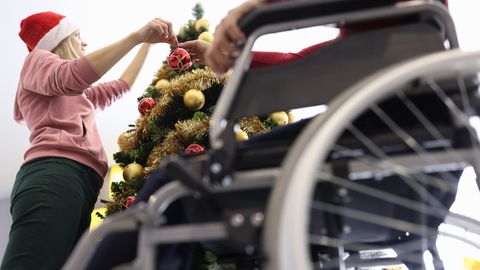 Ein Senior im Rollstuhl sitzt neben einem Weihnachtsbaum, den eine Pflegerin gerade schmückt
