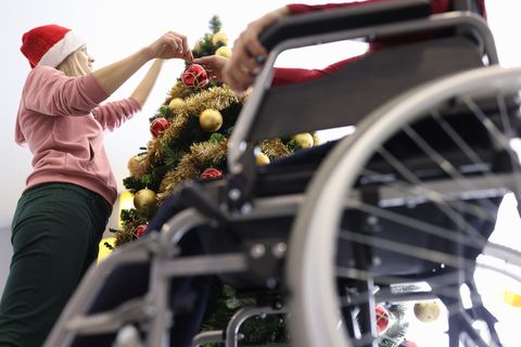 Ein Senior im Rollstuhl sitzt neben einem Weihnachtsbaum, den eine Pflegerin gerade schmückt