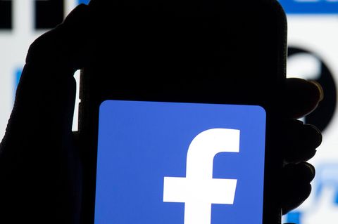 Eine linke Hand hält ein Smartphone, auf dessen Display das blau-weiße Logo von Facebook leuchtet