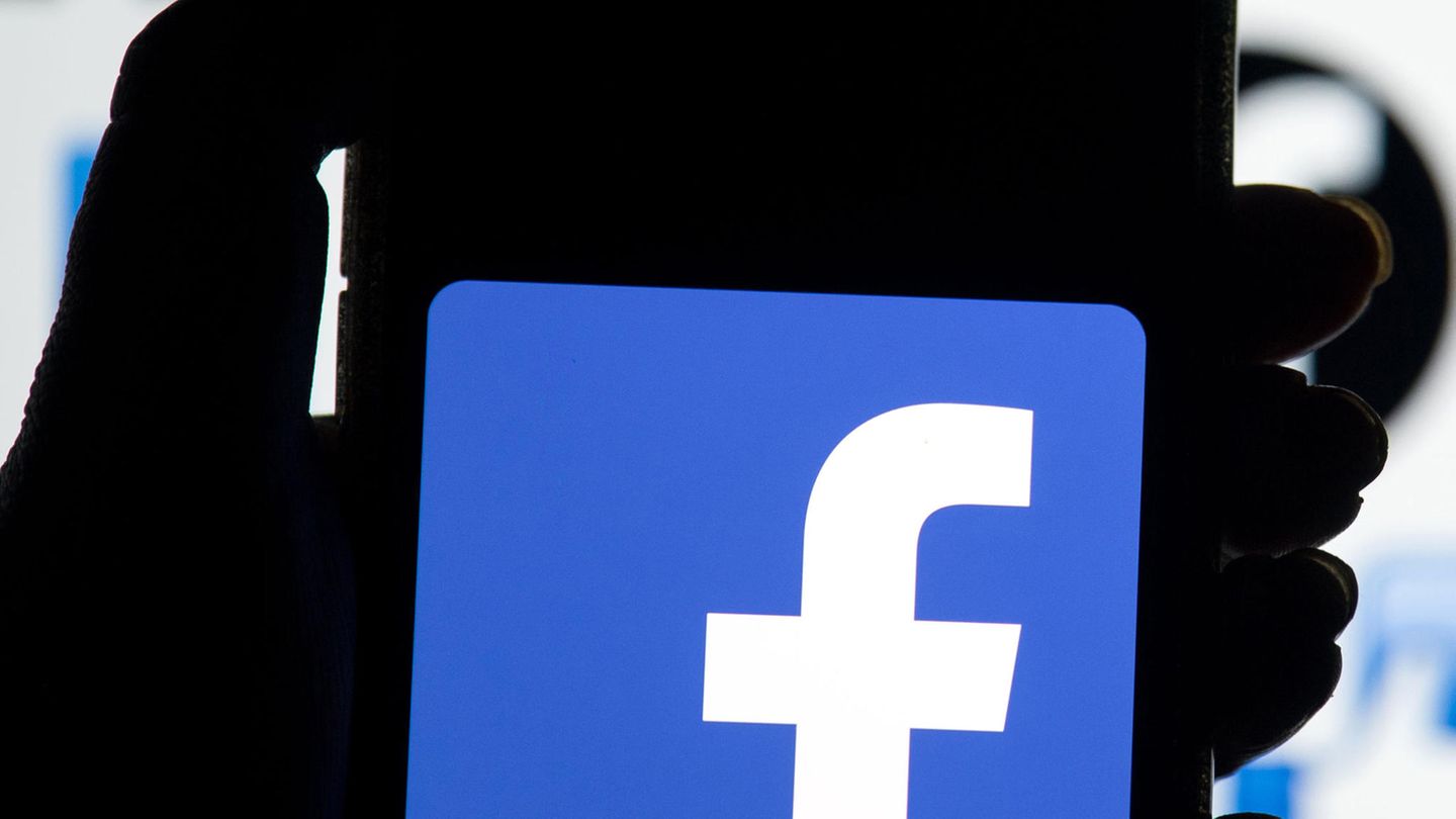 Eine linke Hand hält ein Smartphone, auf dessen Display das blau-weiße Logo von Facebook leuchtet