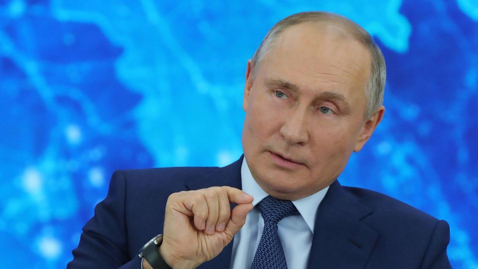 Vor einer blau beleuchteten Weltkarte sitzt Russlands Präsident Wladimir Putin im Anzug und gestikuliert mit der rechten Hand