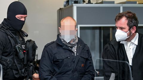 Der Angeklagte von Halle steht im Gerichtssaal zwischen einem vermummten Polizisten und einem Mann iin Robe
