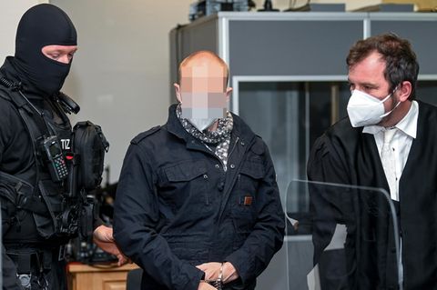 Der Angeklagte von Halle steht im Gerichtssaal zwischen einem vermummten Polizisten und einem Mann iin Robe