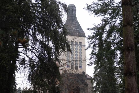 Zwischen Tannen hindurch ist der quadratische und sandfarbene Turm des Krematoriums Zittau zu sehen