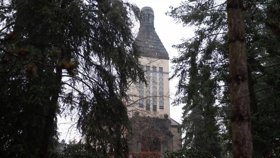 Zwischen Tannen hindurch ist der quadratische und sandfarbene Turm des Krematoriums Zittau zu sehen